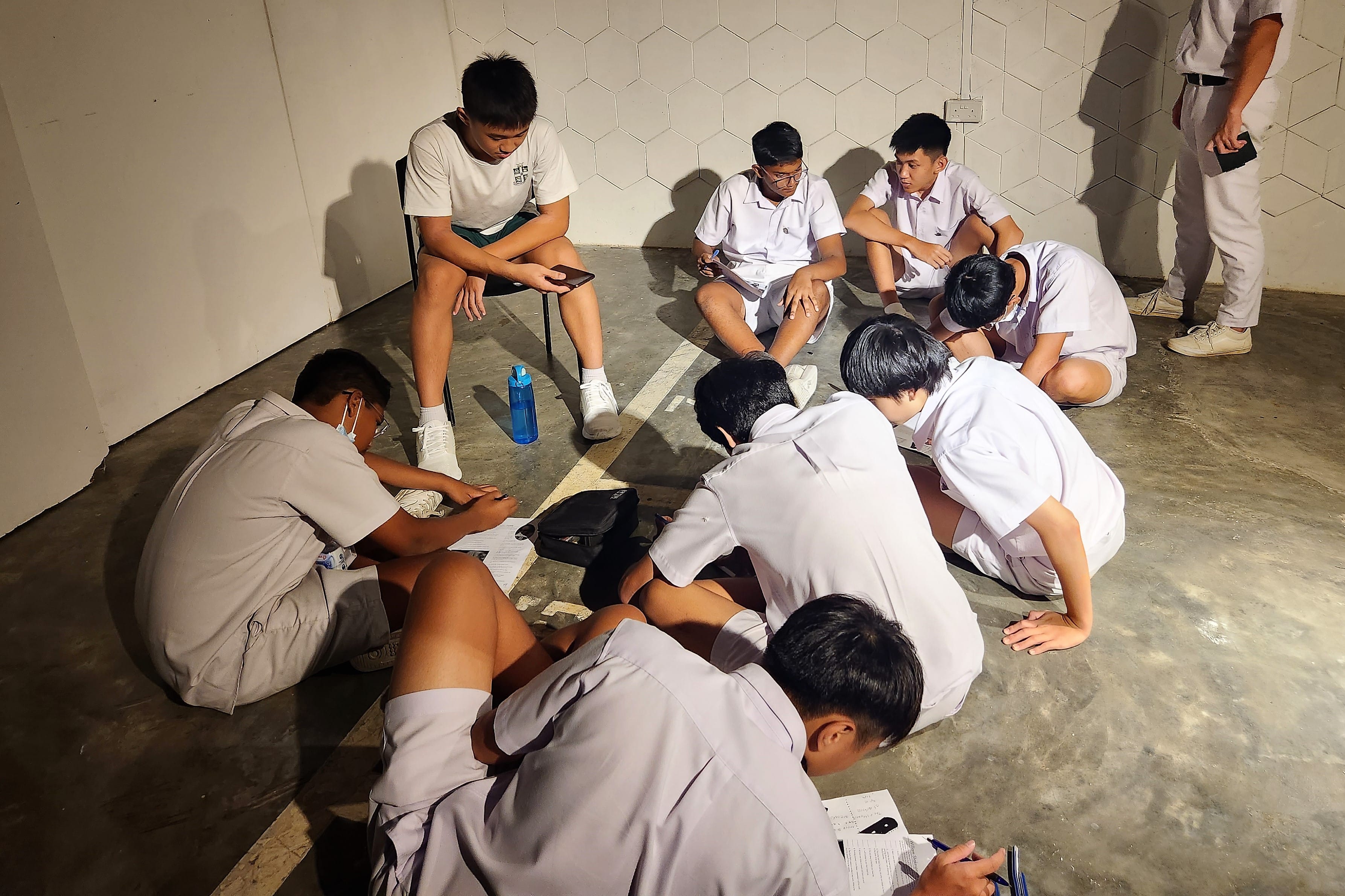 Students sit in a circle on the floor doing a worksheet activity together in a dim indoor hallway.